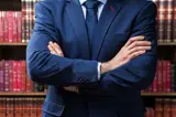 Een man in een blauw pak, armen over elkaar. Boekenkast op de achtergrond.